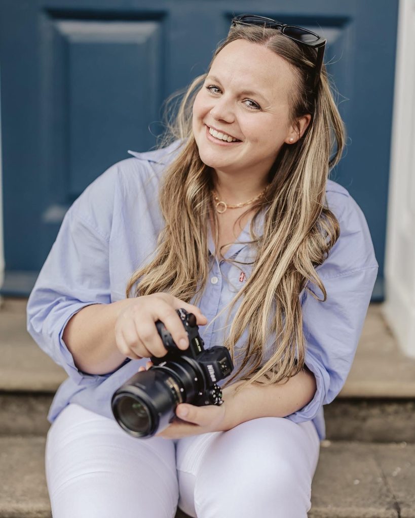 about me page and the photographer wearing a blue top, white jeans and holding a camera whilst looking and smiling at the camera