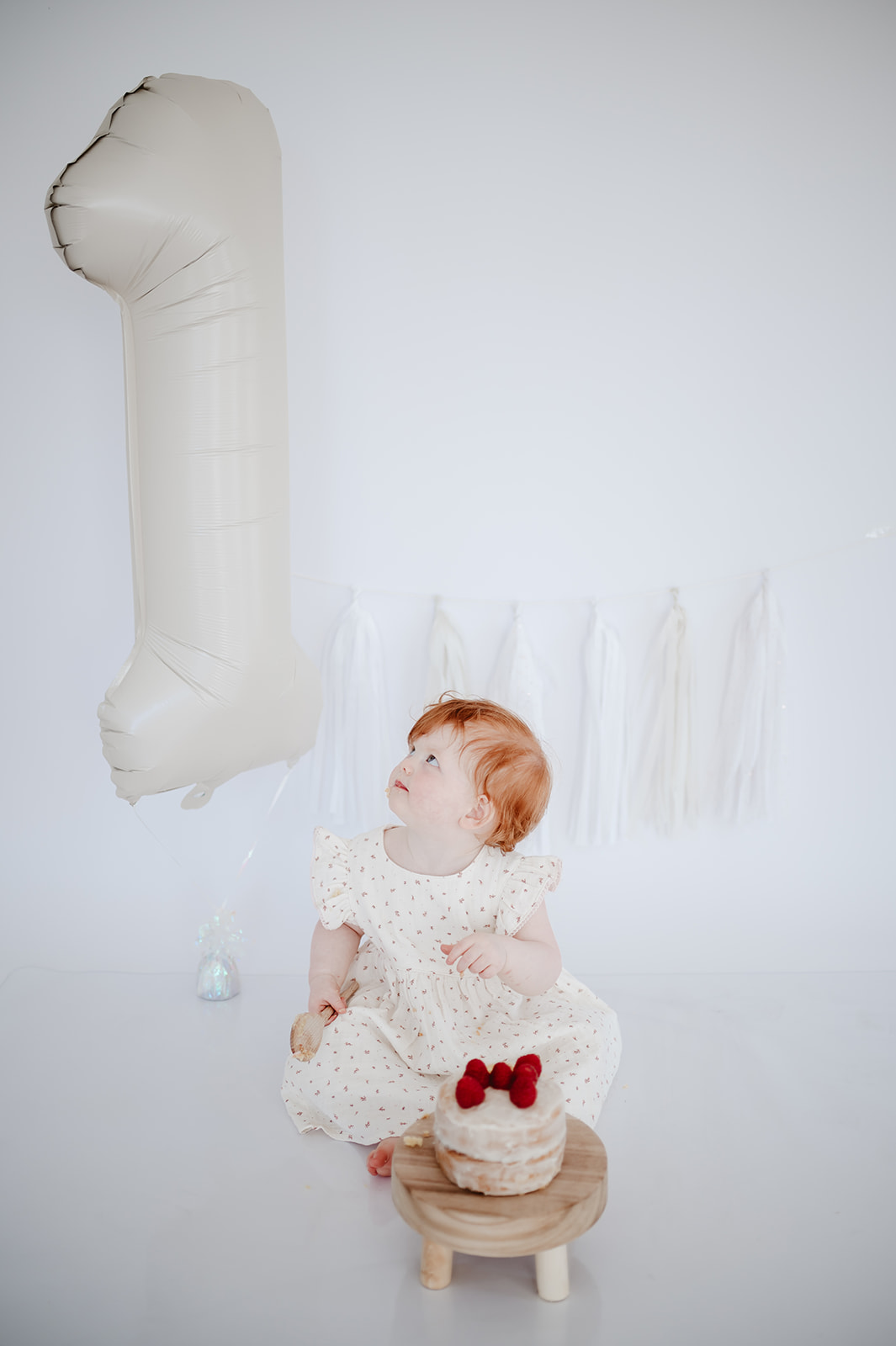 1st balloon with white backdrop and paper banner, with a baby holding a wooden spoon sitting in front of a butter cream cake on a wooden stand.