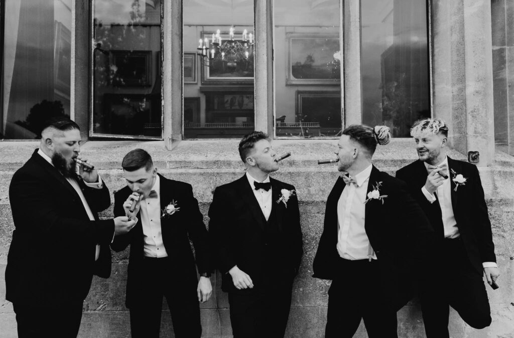 Groomsmen with groom in black tie attire and smoking  cigars against the venue wall in a black and white image