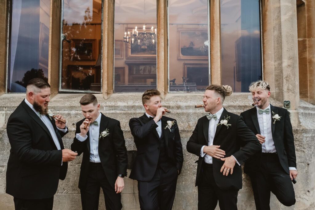 Groom with wedding party in black tie and smoking cigars against the venue wall