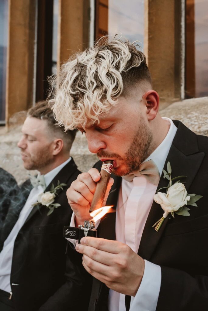 Groomsmen in black tie attire and smoking a cigar against the venue wall