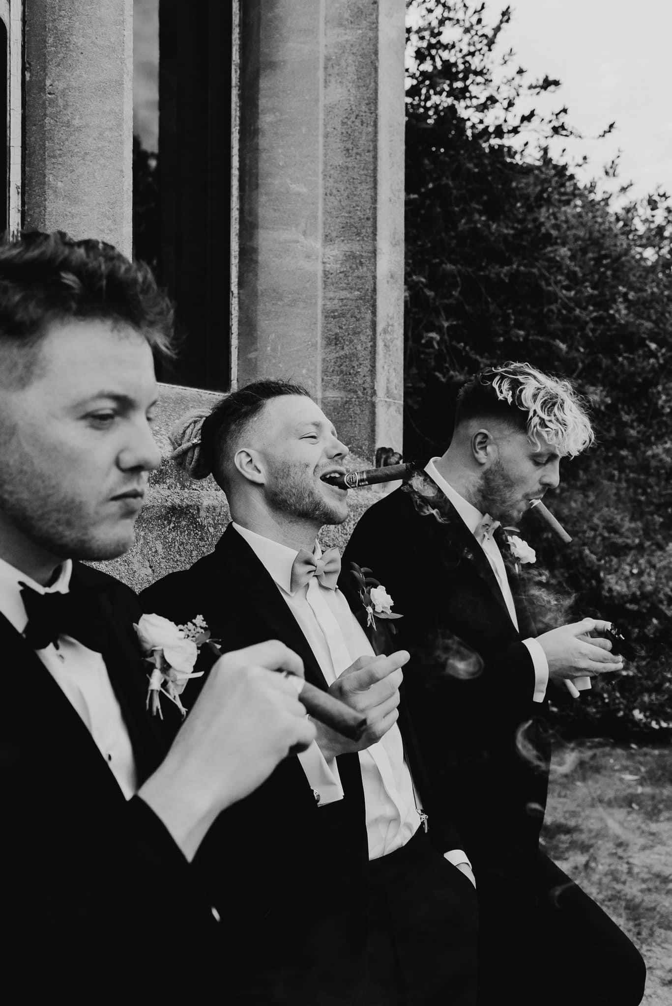 Groom with wedding party in black tie attire and smoking cigars against the venue wall