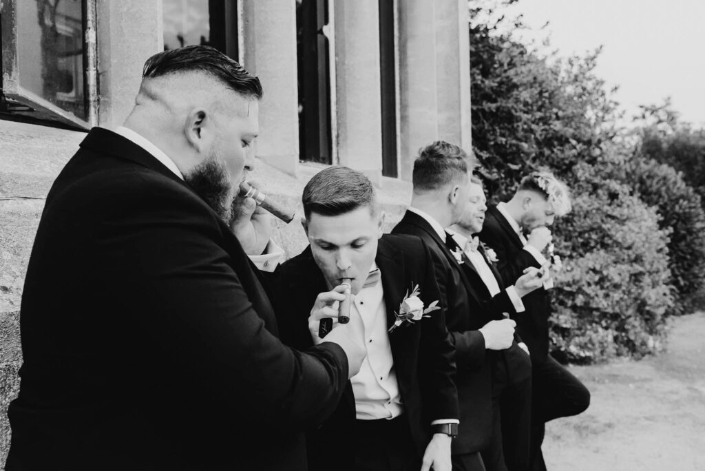 black and white image of Groom with wedding party in black tie and smoking cigars against the venue wall
