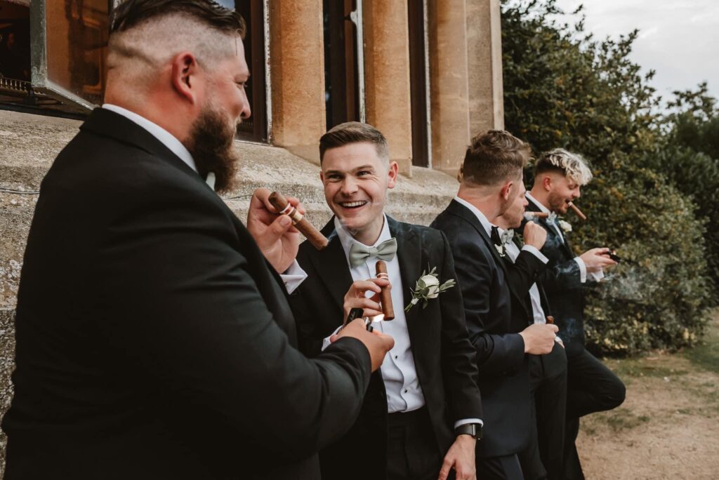 Groom with wedding party in black tie and smoking cigars against the venue wall