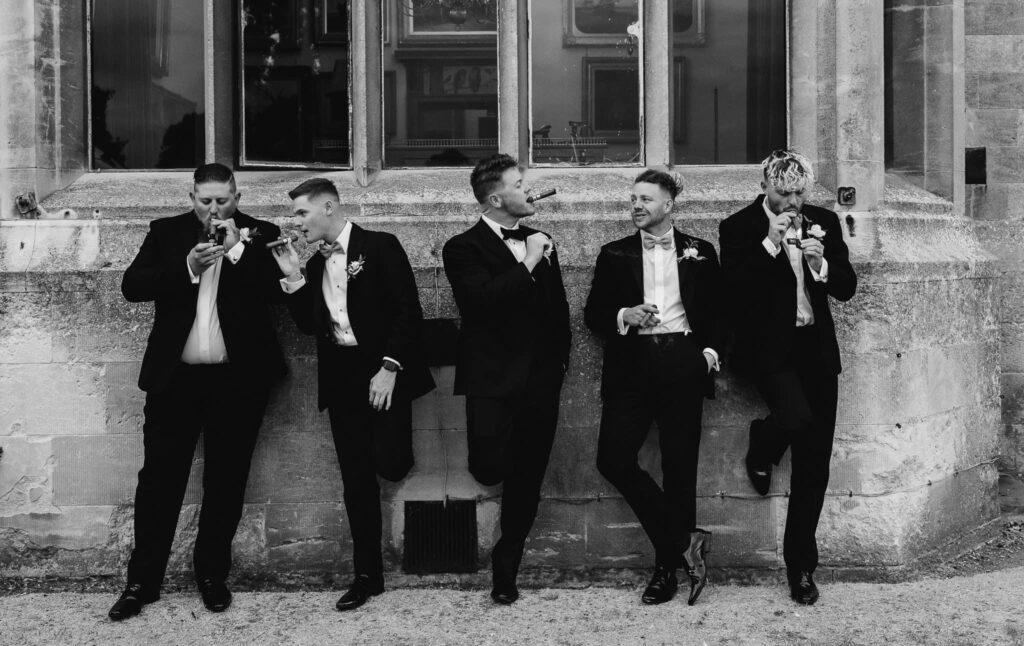 black and white image of groom with wedding party in black tie and smoking cigars against the venue wall