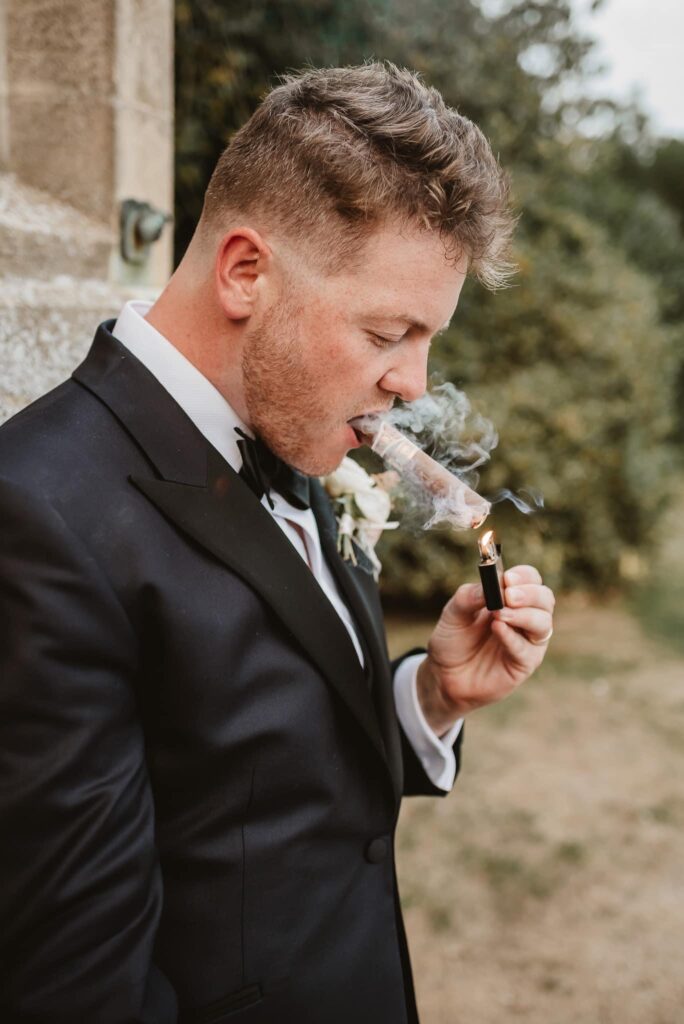 Groom in black tie attire and smoking a cigar against the venue wall