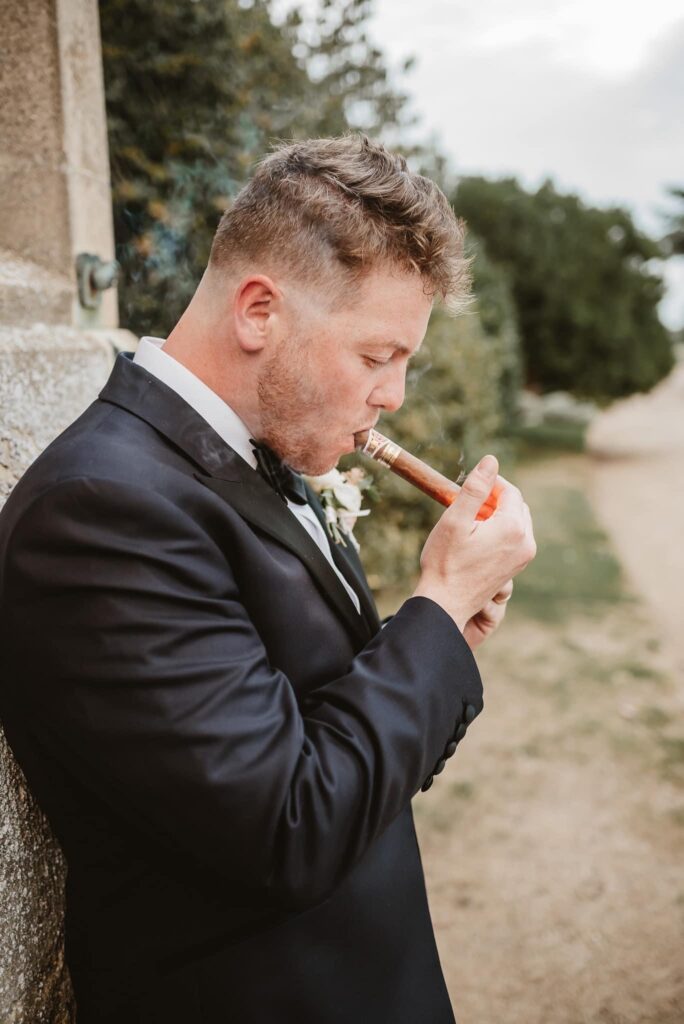 Groom in black tie attire and smoking a cigar against the venue wall