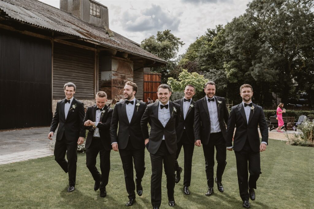 groom dressed in black tie walking with groomsmen outside the venue of the manor barn cambridge, all laughing and smiling. 