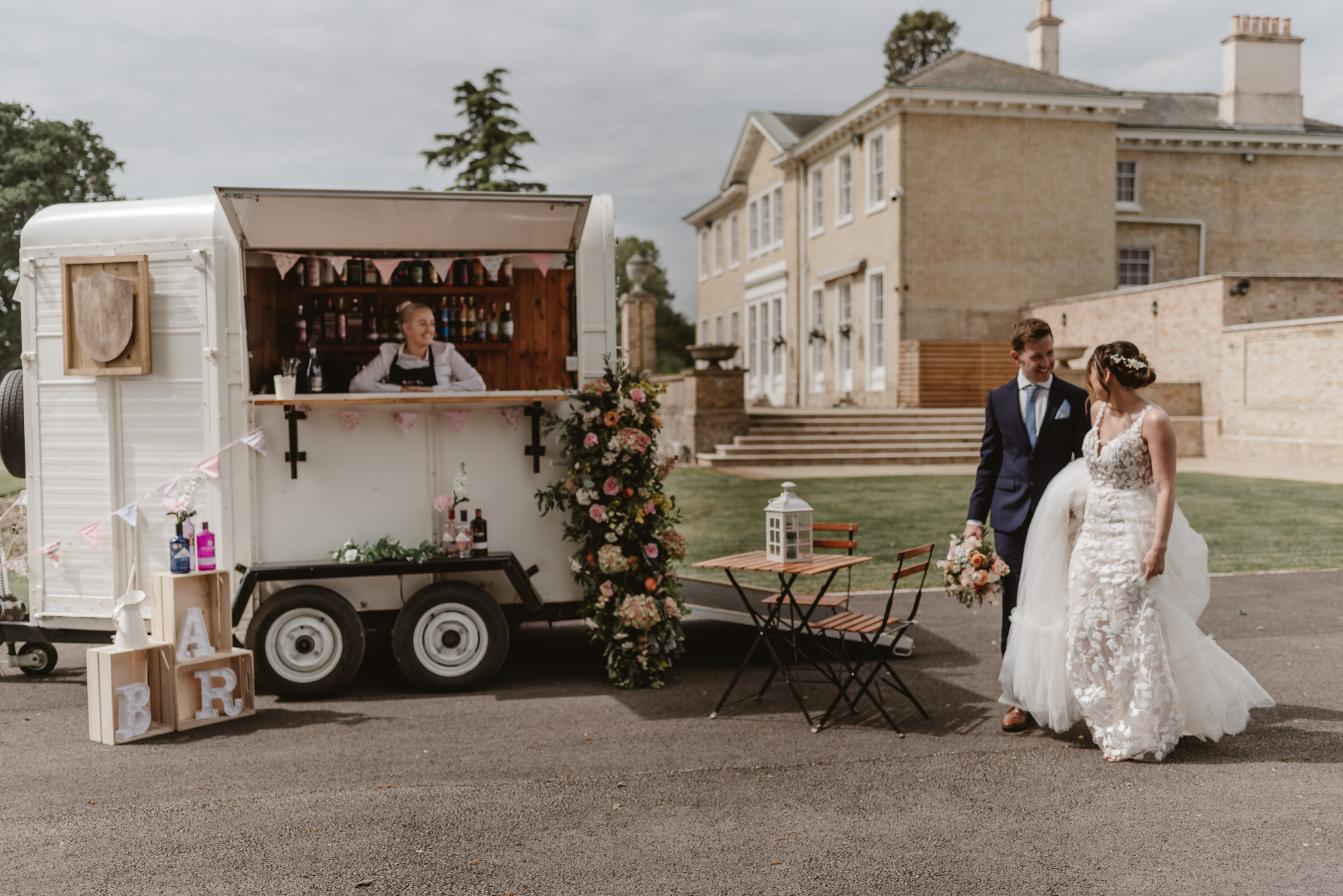 Remote bar decorated in flowers with a bride and groom walking towards it with the wedding venue in background