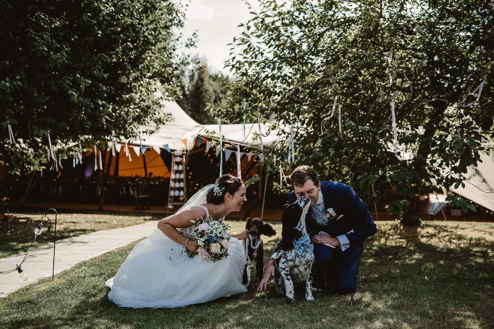 bride and groom holding their dogs facing each other sitting down on the grass smiling outside their wedding venue