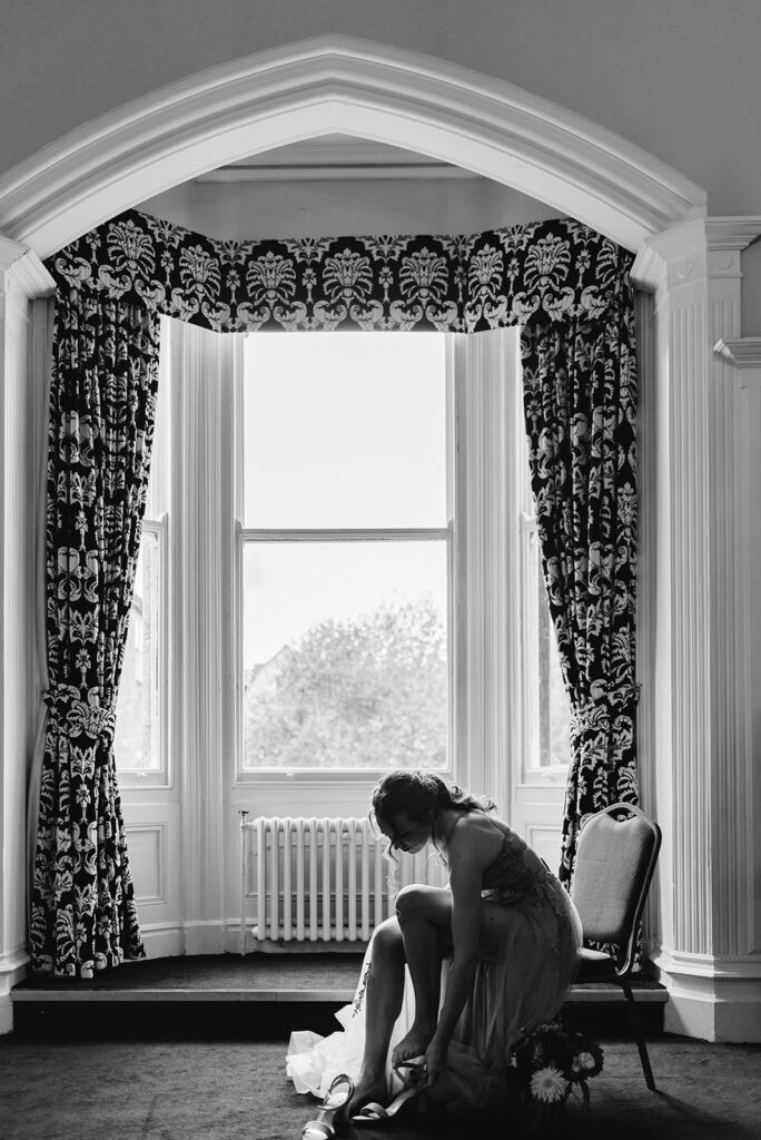 Bride sitting down and putting on shoes in front of window in a black and white image