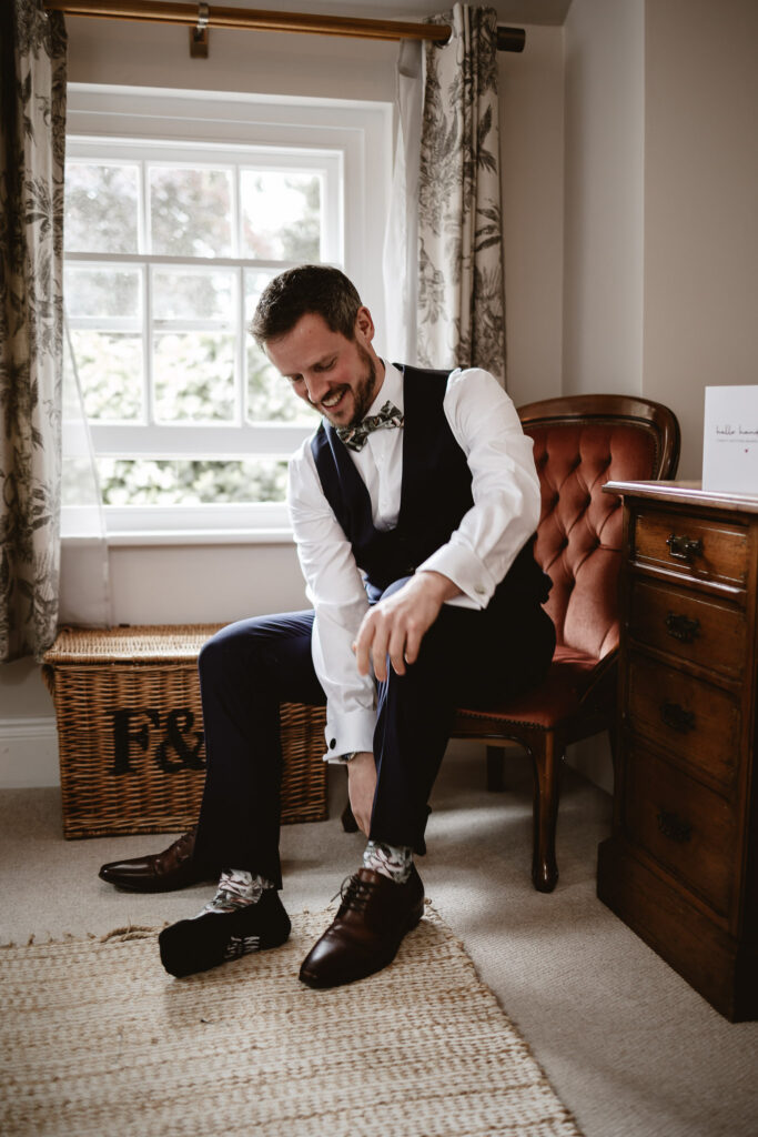 groom putting on his wedding shoes in his wedding attire sitting in a chair in the honey moon suite at the wedding venue