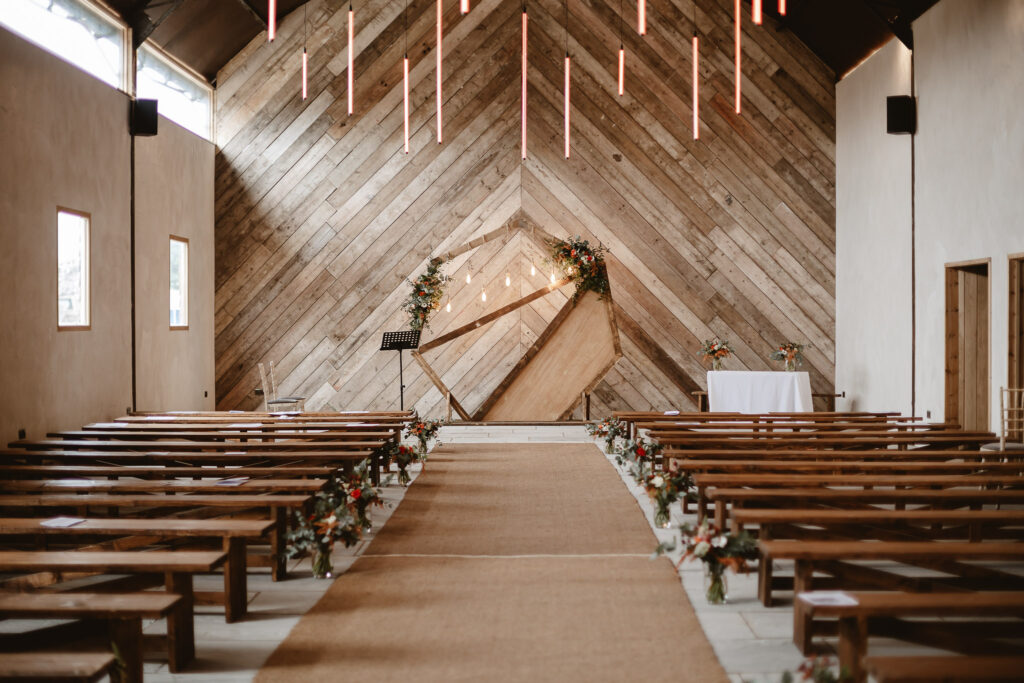 The manor barn ceremony room with a wooden feature wall, cream walls, wooden frame work with wooden benches, low hanging neon lights and a flowery wooden with lights center piece.