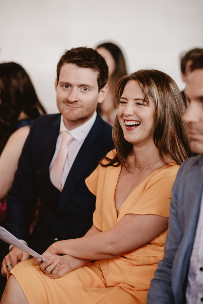 male dressed in suit pulling a face at the camera while female in a orange dress is laughing 