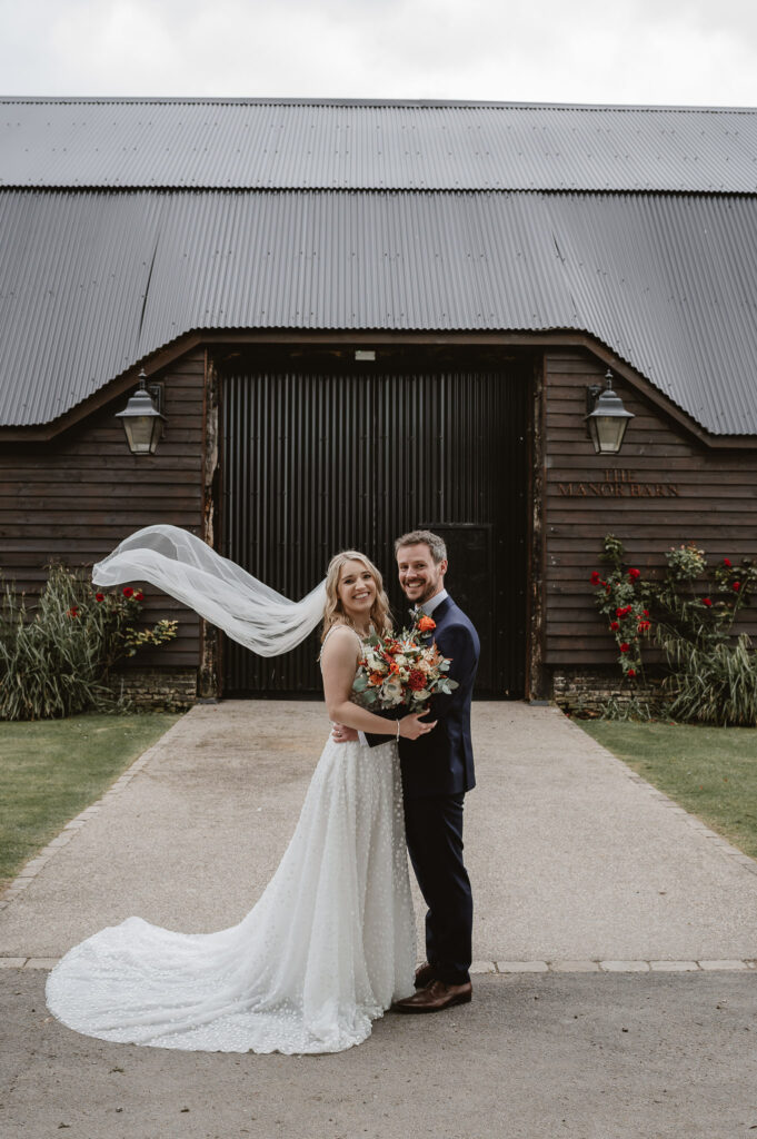 bride and groom standing together smiling looking at the camera and holding flowers with the brides vail blowing in the wind