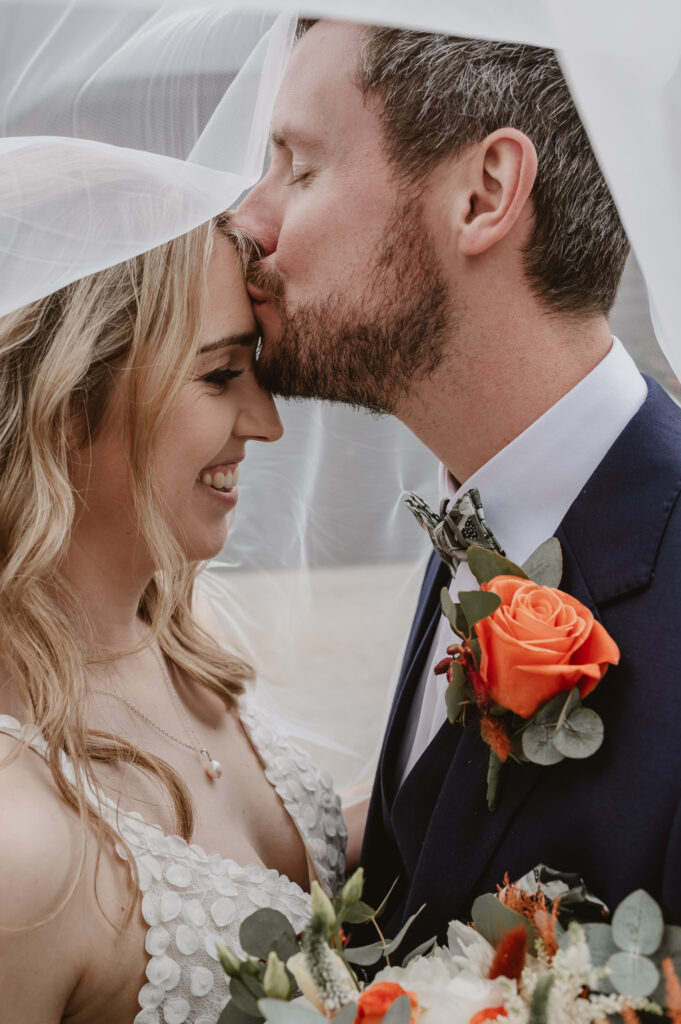 bride and groom under vail, and groom kissing the bride on the forehead underneath the vail