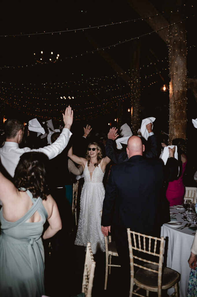 guests throwing napkins in the air while bride high fives the guests whilst wearing glasses