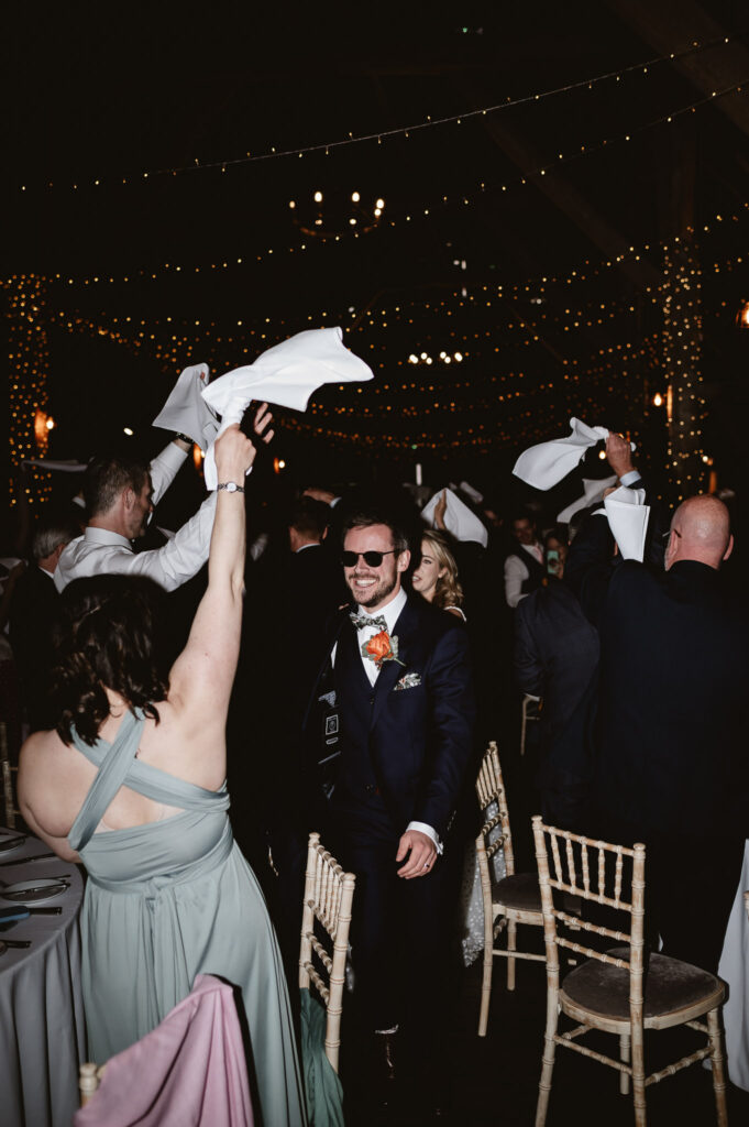 guests throwing napkins in the air while groom is smiling whilst wearing glasses