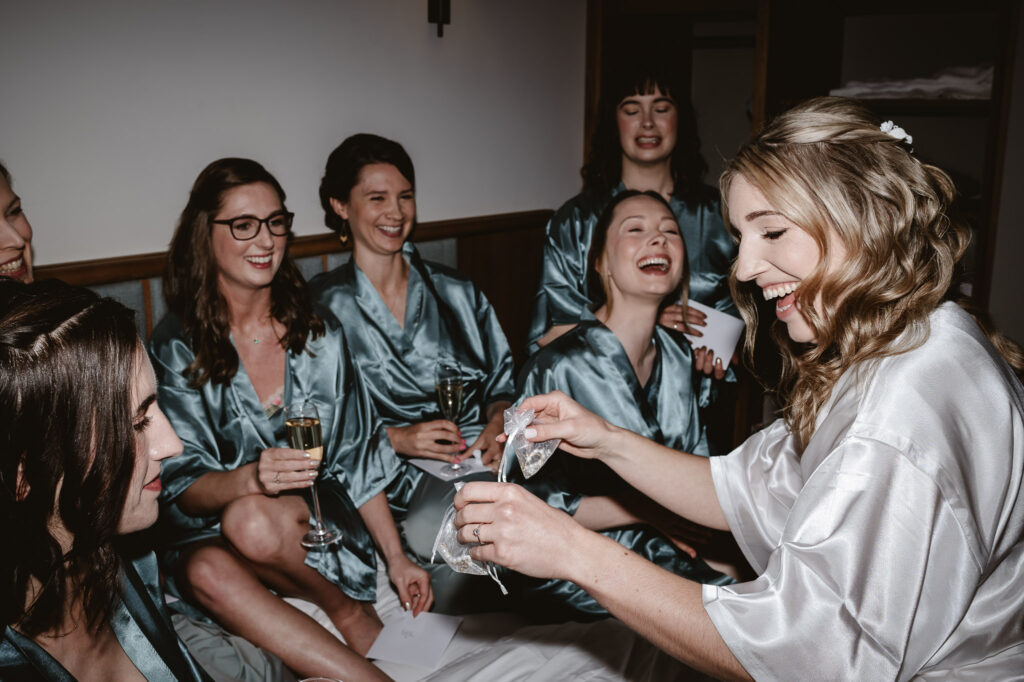 bridesmaids and bride in silk wedding robes drinking bubbly sitting on the bed laughing and the bride is handing out gifts. 