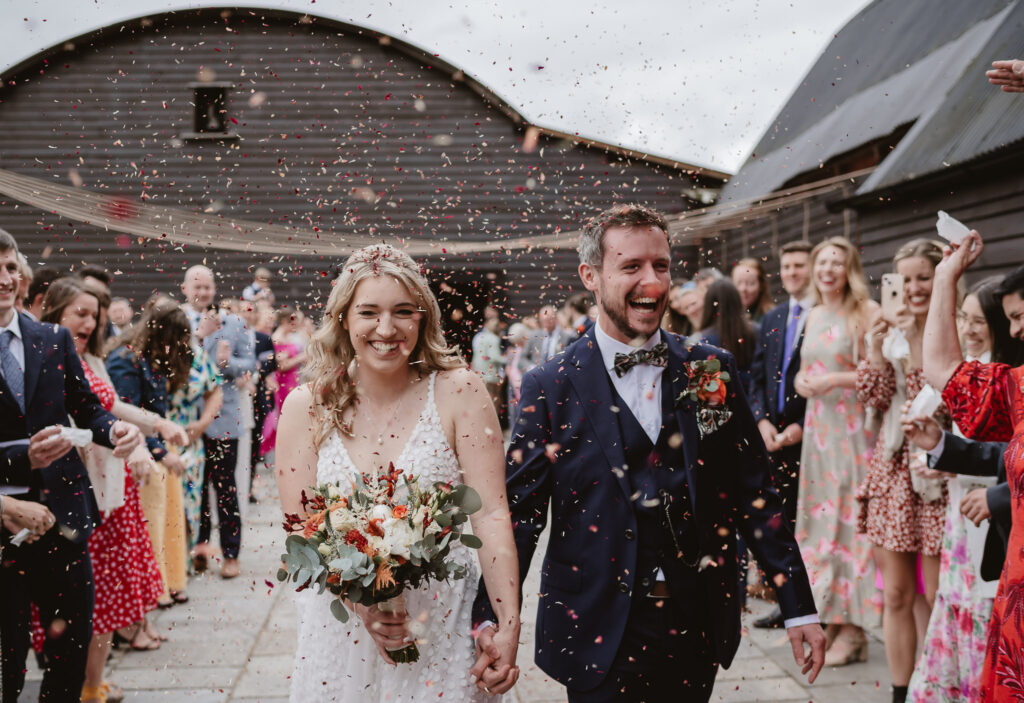 bride and groom holding hands and smiling walking through confetti outside with wedding guests eitherside