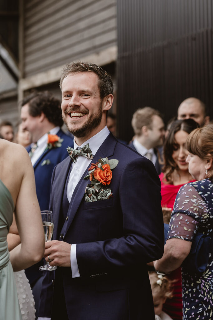 Groom smiling holding a glass of bubbly in a blue suit with a orange rose button hole and bow tie