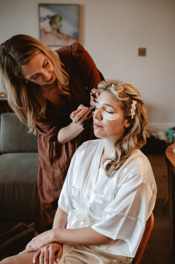 make up artist getting the bride ready for her wedding day whilst the bride relaxes in her white silk robe and completes her wedding skin care routine