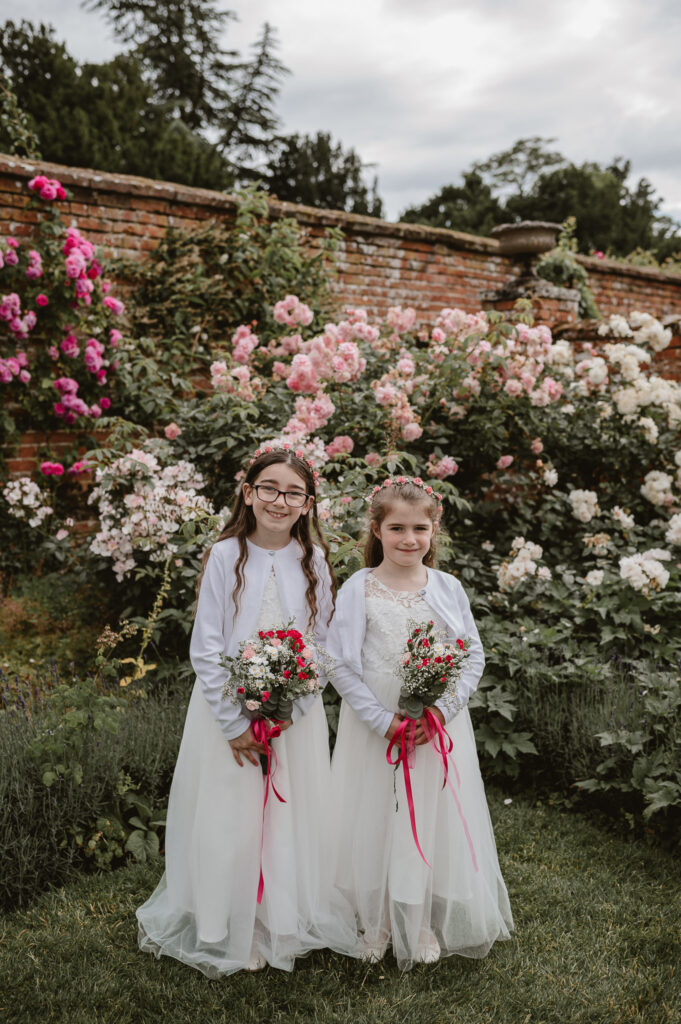 two flower girls dressed in white, with flower crowns and holding flowers standing next to each other smiling in the rose garden at a wedding venue. 