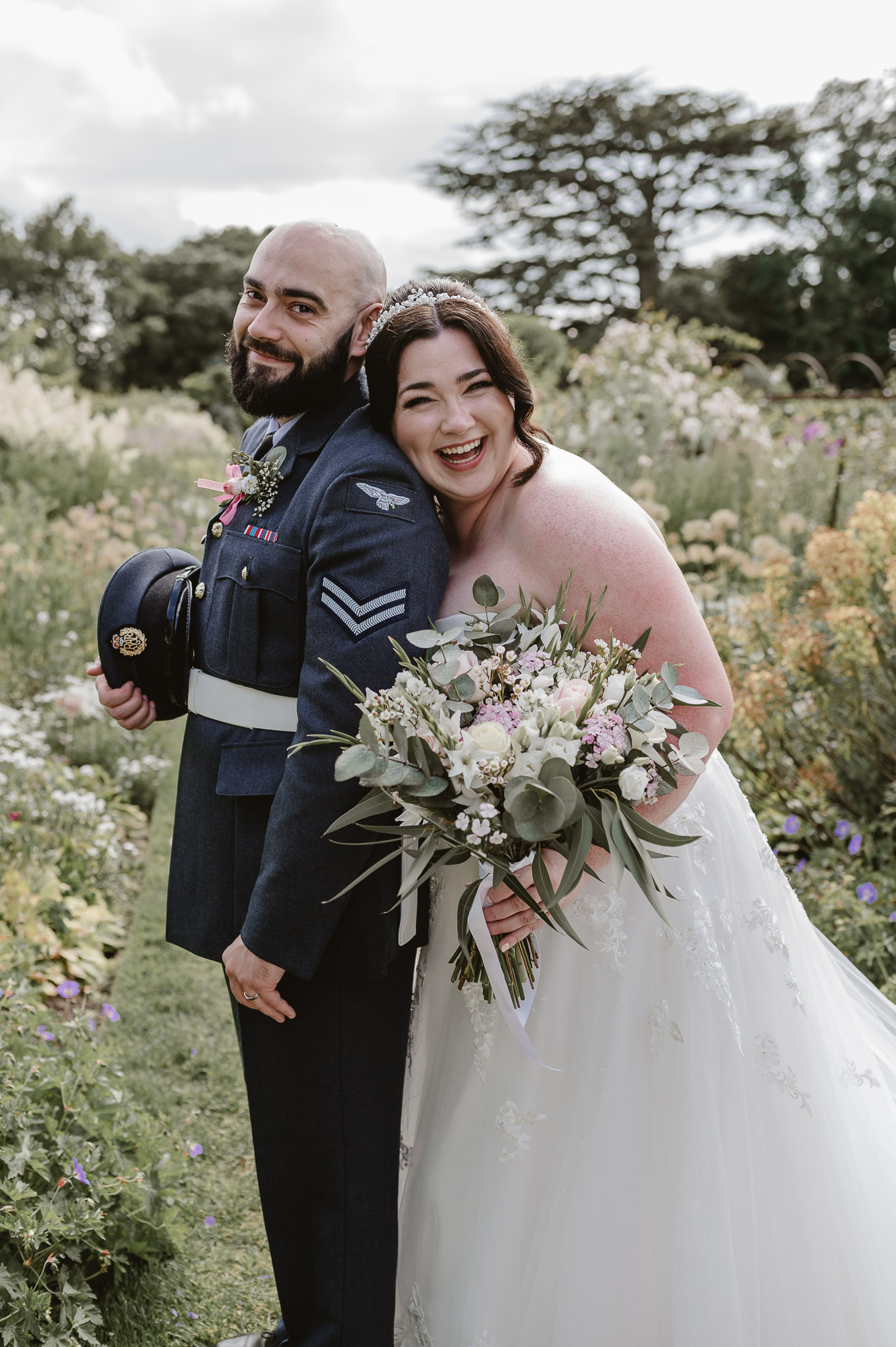 Groom dressed in RAF uniform with bride leaning again his back holding flowers while they both look at the camera and smile in a flower garden