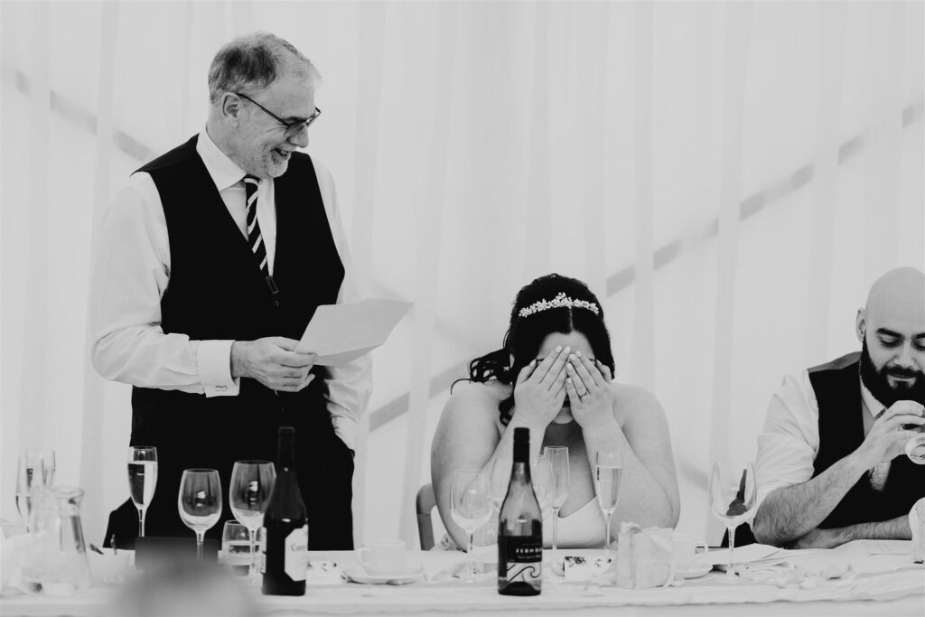 black and white image of wedding speeches with the father of the bride laughing whilst holding paper and looking down at his daughter, the bride with her hands covering her face.