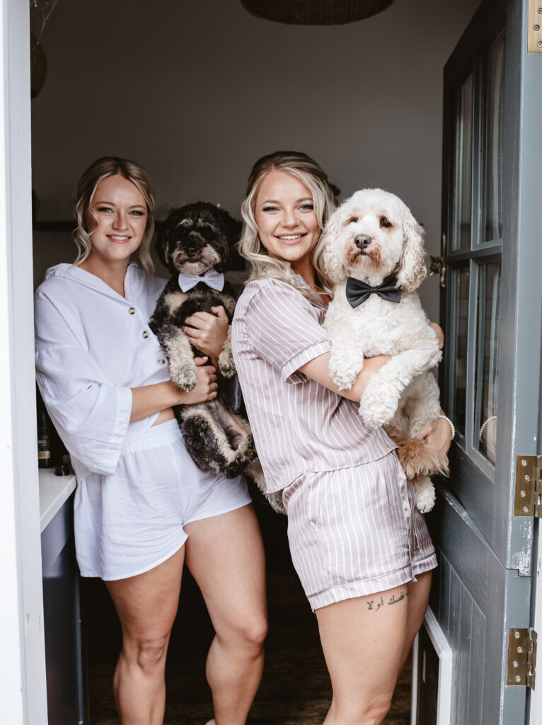 bride and bridesmaid in their pjs holding their dogs that are wearing a bow ties standing in the doorway during bridal prep at the wedding venue