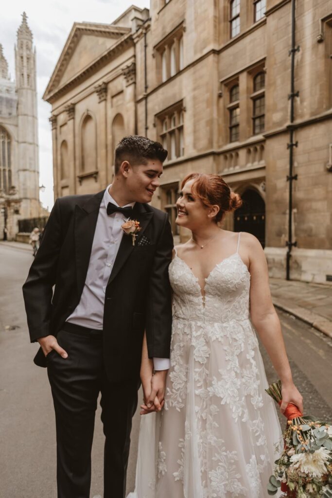 bride and groom holding hands walking and smiling looking at each other in the middle of Cambridge city